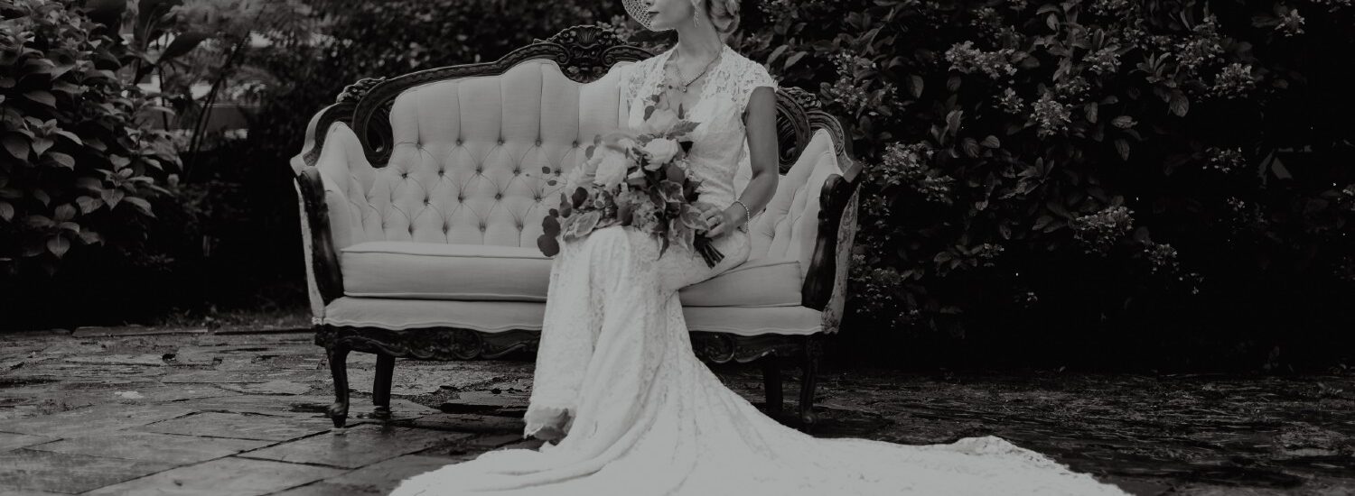 Black and white photograph of a bride in white with a bouquet sitting on an antique loveseat outdoors